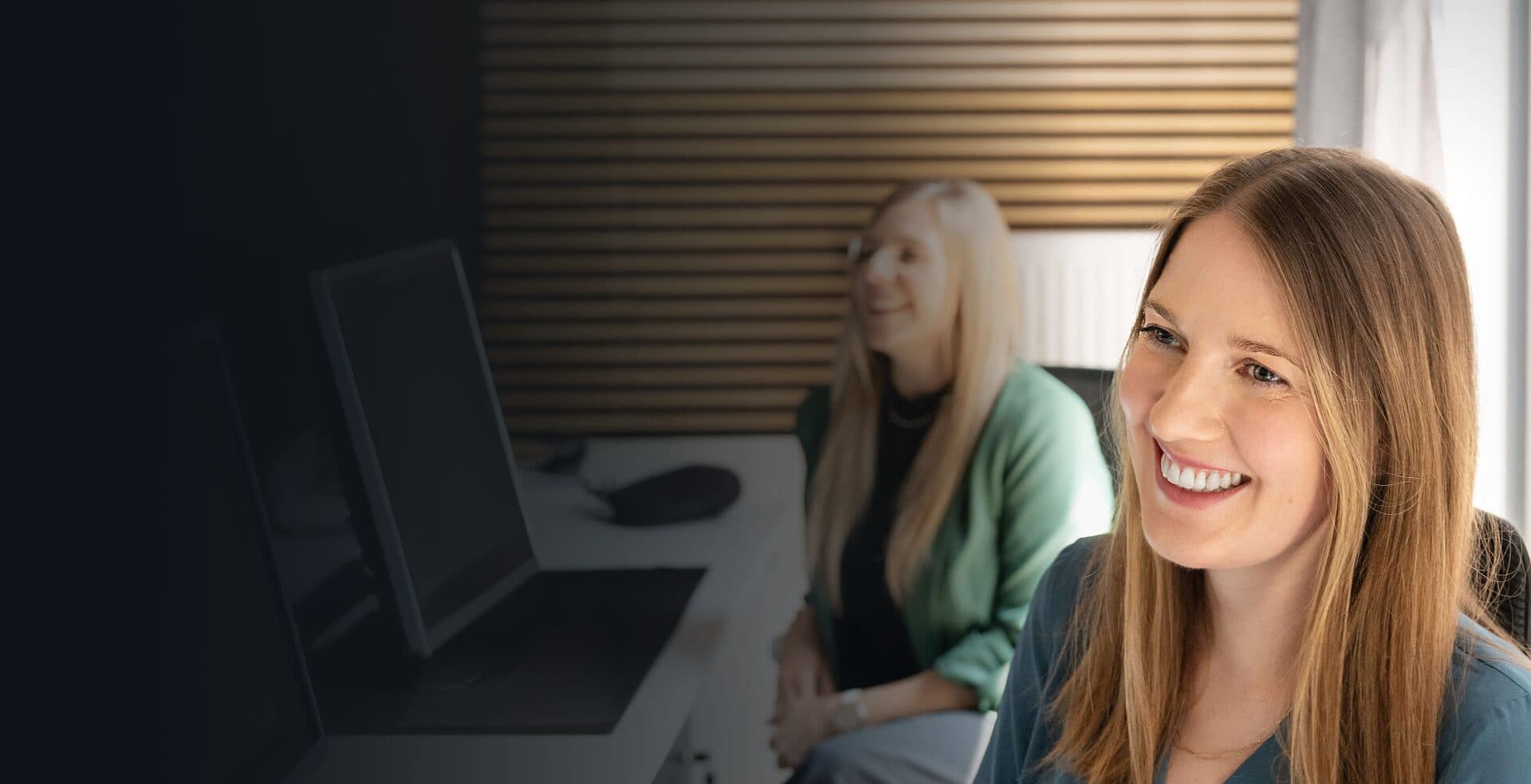 Lächelnde Mitarbeiterin am Arbeitsplatz, im Hintergrund eine Kollegin am Schreibtisch