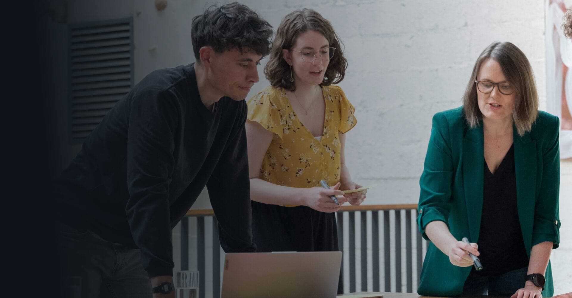 Three colleagues at a table with a laptop and sticky notes talking through their ideas