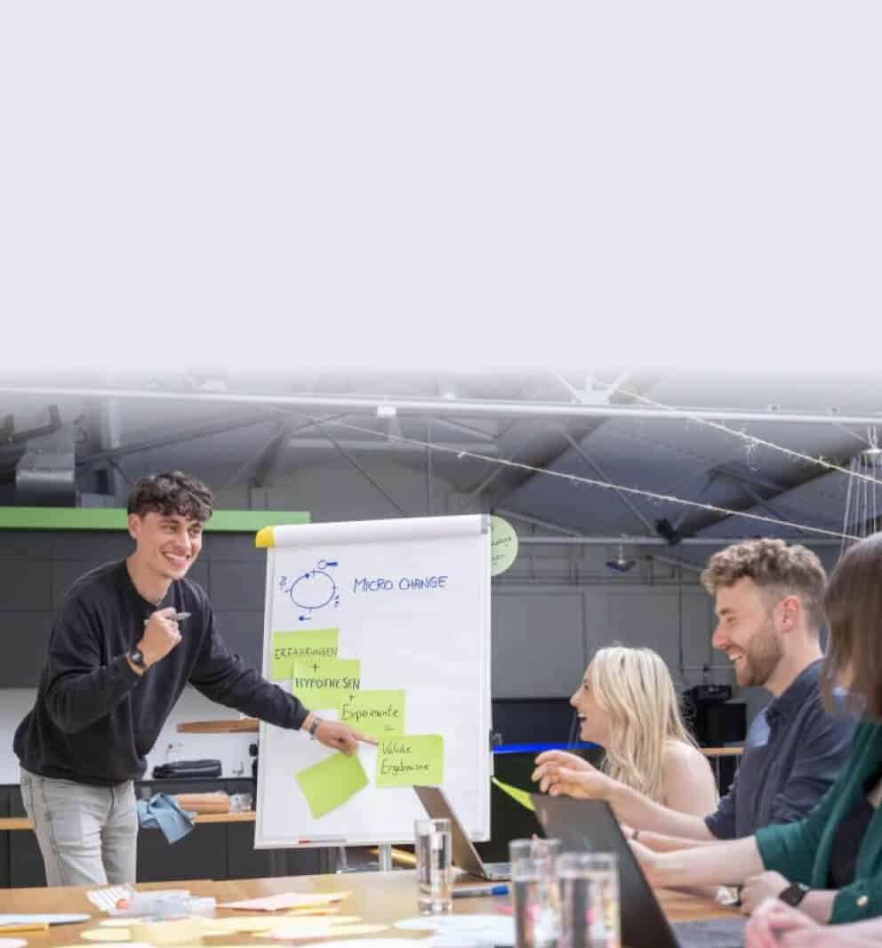 Young man presenting a “Micro Change” flipchart with sticky notes to a laughing team