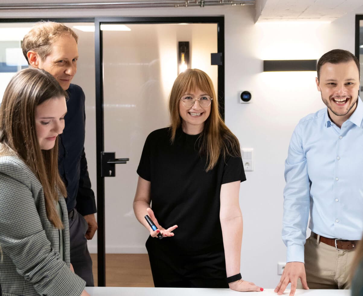 Four team members standing at a meeting table in good spirits, one colleague facilitating with a pen in hand