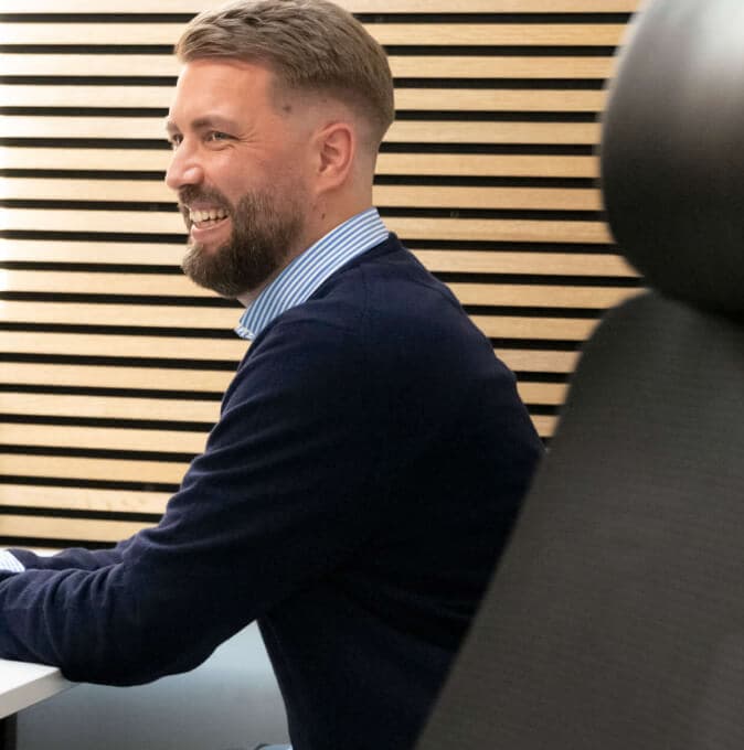 Lachender Mitarbeiter an seinem Schreibtisch vor einer Holzlamellenwand
