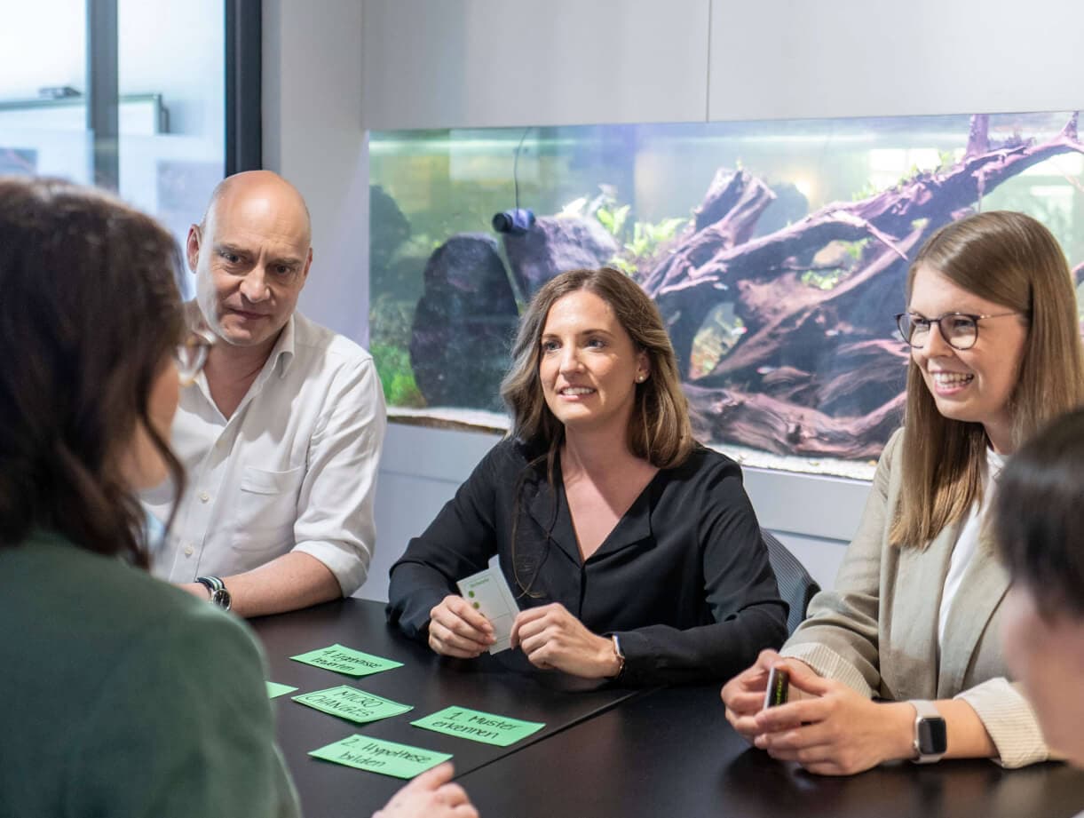Workshop group at a table with green cards showing steps such as “Recognise patterns” and “Form a hypothesis”