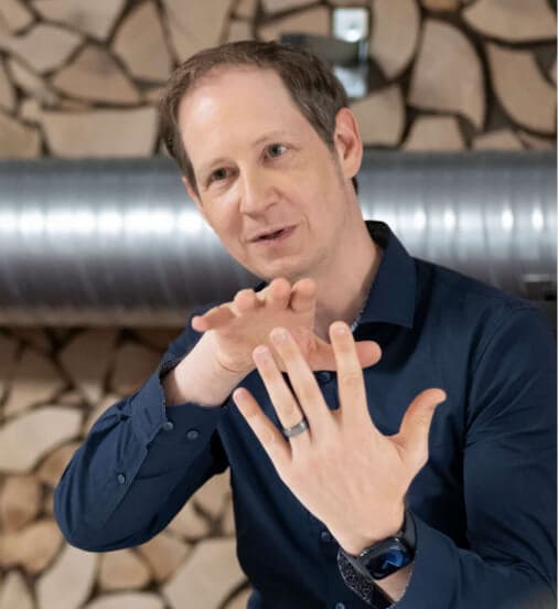 Consultant explaining with both hands, a wall of stacked logs behind him