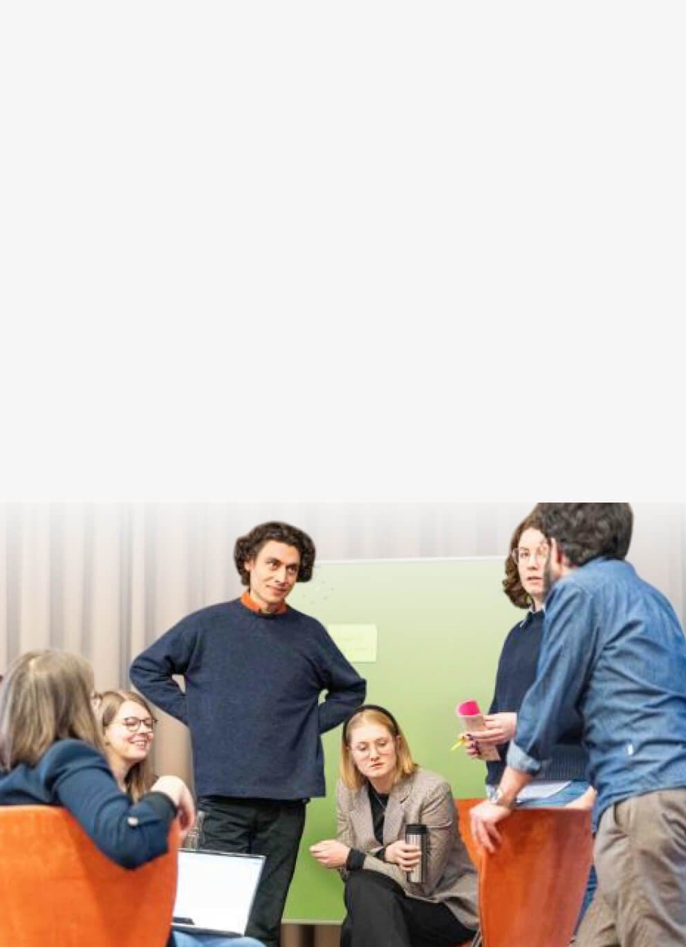 Workshop group in discussion, some seated and some standing, in front of a green pinboard