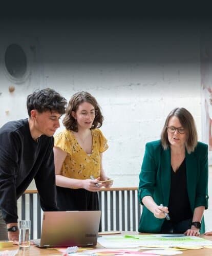 Three colleagues at a table with a laptop and sticky notes talking through their ideas