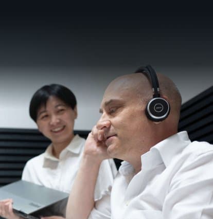 Mann mit Headset telefoniert, eine Kollegin mit Laptop verfolgt das Gespräch lächelnd